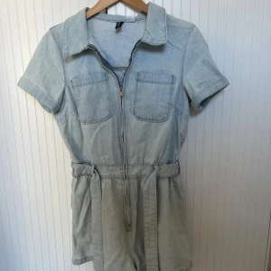 Ljusblå jeans playsuit från H&M Divided - Snygg ljusblå jeans playsuit från H&M Divided med korta ärmar, krage och två bröstfickor. Dragkedja framtill och tillhörande bälte i midjan.