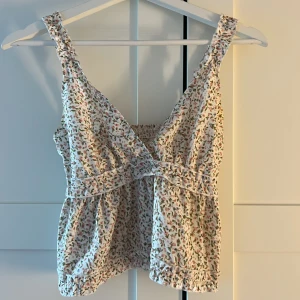 Blommig linnetopp med volangdetaljer - Supersöt linnetopp med småblommigt mönster i rosa, grönt och beige på vit botten. Toppen har breda axelband, v-ringning och volangdetaljer längs kanterna. Smockad rygg för extra komfort och luftig passform. Perfekt för varma dagar och somriga vibes.