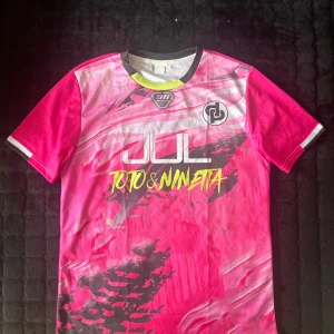 Jul T-shirt Toto & Ninetta - Snygg Jul tröja i rosa med coolt grafiskt mönster och tryck från Toto & Ninetta. Framsidan har logga och text i vitt och gult, baksidan har nummer 7 och Ninetta.