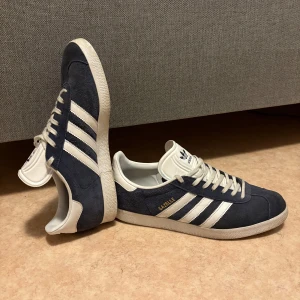 Adidas Gazelle blå sneakers strl 42 - Säljer ett par klassiska Adidas Gazelle sneakers i marinblå mocka med vita detaljer och de ikoniska tre ränderna på sidan. Skorna har snörning, vit platt sula med hexagonmönster och rund tå. Guldtexten 'GAZELLE' på sidan ger extra stilpoäng. Perfekt för dig som gillar retrovibe.
