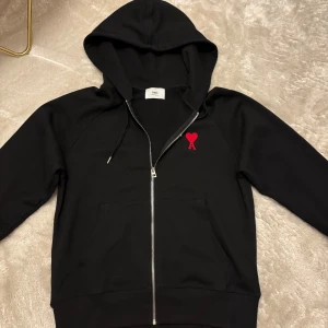 NY Ami hoodie med rött hjärta - Svart hoodie från Ami Alexandre Mattiussi i storlek M. Tröjan har dragkedja, huva med snörning och en stor ficka framtill. På bröstet finns ett broderat rött hjärta med bokstaven A. Hoodien är knappt använd och i stort sätt ny. Påsen tillkommer! Hör av er vid frågor 