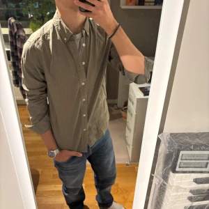 Olivgrön skjorta med knappar - Säljer en stilren olivgrön skjorta med klassisk krage och vita knappar hela vägen. Skjortan har lång ärm och en normal passform som funkar till jeans eller chinos. Perfekt för dig som gillar enkel och clean stil.