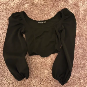 Svart croppad topp PrettyLittleThing - Säljer en svart croppad topp från PrettyLittleThing med långa puffärmar och rundad urringning. Toppen har en figurnära passform och är perfekt för dig som gillar trendiga och stilrena plagg. Materialet känns mjukt och följsamt.