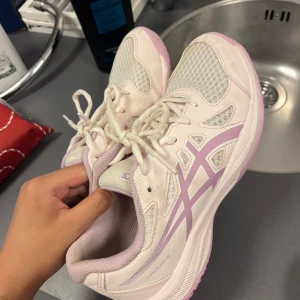 Ljusrosa / lila Asics sneakers - Säljer ett par ljusrosa / ljuslila Asics sneakers med vita och lila detaljer. Skorna har ventilerande meshpartier, klassisk snörning och en platt sula. Perfekta för dig som gillar sportig stil och vill ha bekväma skor med snygg design. HELT NYA!! Köpt för runt 900kr