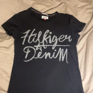Svart Hilfiger Denim t-shirt XS - Svart t-shirt från Hilfiger Denim i storlek XS med stort tryck i grå på bröstet. Klassisk rund halsringning och korta ärmar. Perfekt för dig som gillar en enkel men snygg look med tydlig logga framtill.