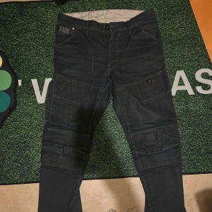 Svarta jeans från G-Star RAW - Säljer ett par svarta jeans  från G-Star RAW, modell R33/01. Byxorna har flera fickor med knappstängning, coola sömmar och detaljer både fram och bak. Materialet är slitstark jeans och passformen är loose med tapered ben. Perfekt för dig som gillar streetwear och funktionella plagg.