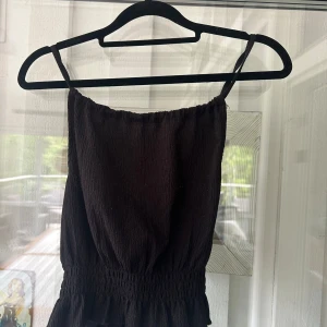 Svart linne med kors i ryggen från Gina Tricot - Supersöt svart linne från Gina Tricot med smala axelband och smock i midjan. Linnet har crinklat tyg och ett kors i ryggen. Storlek S (Toppen är använd men i fint skick)