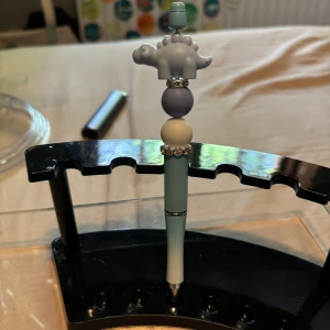 Penna - Bilderna visar en blå och vit penna dekorerad med pärlor och en söt dinosauriefigur på toppen. Pennan står i ett svart ställ och har en lekfull, trendig design som passar perfekt för dig som vill sticka ut i skolan eller på jobbet. Perfekt accessoar för unga kreativa själar.