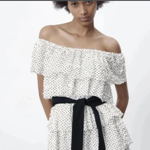 Prickig offshoulder kortklänning med volang - Superfin vit kortklänning med svarta prickar, offshoulder-modell och volangdetaljer. Klänningen har ett brett svart knytband i midjan som ger en snygg siluett. Perfekt för sommarens alla tillfällen!