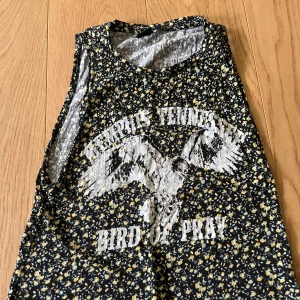 Blommig ärmlös topp från Gina Tricot - Snygg ärmlös topp från Gina Tricot i svart med små gula och vita blommor. Tryck framtill med texten 'Memphis Tennessee Bird of Pray' och en stor örn. Perfekt för dig som gillar statement-plagg och coola tryck.