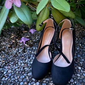 Svarta pumps från Anna Field - Snygga svarta pumps från Anna Field med rund tå, hög klack och smala remmar som korsas över foten och spänns vid ankeln. Skorna har en stilren look och är gjorda i mocka-liknande material. Perfekta för dig som gillar klassisk men trendig stil.