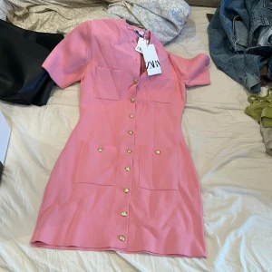 Rosa kortklänning från Zara - Superfin rosa kortklänning från Zara med guldiga knappar framtill och fyra stora fickor. Klänningen har korta ärmar och är gjord i ett strukturerat, stickat material. Perfekt för dig som vill ha en söt och trendig look!
