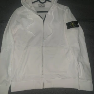 Vit hoodie från Stone Island - Vit hoodie från Stone Island med klassisk logga på vänster ärm. Tröjan har dragkedja framtill, snörning i huvan och stora fickor. Tillverkad i mjuk bomull, perfekt för en clean och stilren look. Tröjan är i ny skick, har inte använt. Inga defekter. Fraktar samma dag. Priset kan diskuteras.