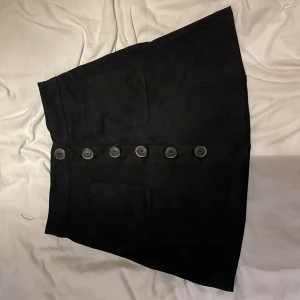 Svart kortkjol med knappar - Svart kortkjol i mockaimitation med stora fickor framtill och snygga knappar längs hela framsidan. Kjolen har en lätt A-linjeformad siluett. 