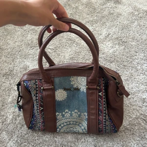 Desigual handväska i skinn och jeans - Säljer en unik handväska från Desigual med bruna skinnpartier, denim i mitten och broderade mönster i färg på sidorna. Väskan har två rejäla handtag, dragkedja upptill och blått mönstrat innerfoder. Perfekt för dig som gillar boho-vibe och detaljer.