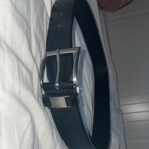 Hugo Boss bälte - Stilrent svart bälte från Hugo Boss i äkta skinn med klassiskt silverfärgat spänne. Bältet har en metallbricka med Boss-loggan och en enkel, rak design som passar till jeans eller kostymbyxor. Perfekt accessoar för en clean och snygg look.