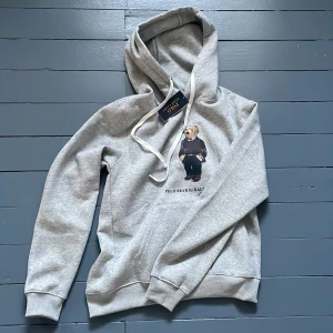 Grå hoodie Polo Bear Ralph Lauren - Snygg ljusgrå hoodie från Polo Ralph Lauren med ikoniska Polo Bear-trycket på bröstet. Tröjan har huva med vita snören och klassisk känguruficka. Perfekt för dig som gillar streetstyle och vill ha något bekvämt men ändå stilrent.