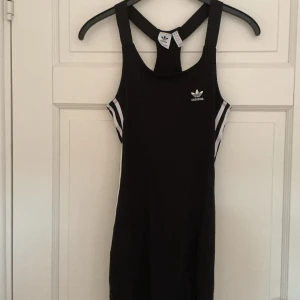 Svart sportig klänning från Adidas - Svart kort klänning från Adidas med klassiska vita ränder på sidorna och broderad logga framtill. Tight passform, ärmlös med brottarrygg och sportig vibe. Perfekt för dig som gillar streetstyle och athleisure. Materialet är mjukt och stretchigt, troligen bomull och polyester.