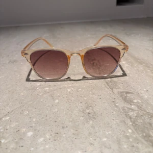 Transparenta solglasögon med bruna glas - Snygga solglasögon med transparent, lätt guldfärgad båge och fyrkantig form. Glasen är bruna med en lätt toning som ger en cool vibe. Perfekta för soliga dagar och en stilren look. Bågarna är i plast och känns lätta på ansiktet.