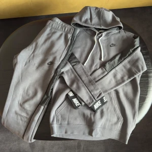 Grå Nike mjukisset - Säljer en grå mjukisset från Nike med matchande hoodie och byxor. Hoodien har huva med vit dragsko, svarta detaljer på ärmarna och Nike-logga på bröstet. Byxorna har resår i midjan och Nike-logga på benet. Perfekt för chill eller träning.