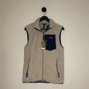 Patagonia Fleece Väst  - Säljer en beige fleeceväst från Patagonia i storlek S. Västen har blå detaljer, hög krage, dragkedja framtill och en blå bröstficka med dragkedja. Perfekt för lager-på-lager och chill outdoor-vibe. Mjuk och skön med två sidofickor med dragkedja. Unisex storlek så sitter smått mindre. Tags och kvittot finns kvar! Hör gärna av er vid flera frågor eller funderingar! 