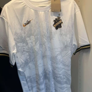 Vit AIK fotbollströja från Nike - Snygg vit AIK fotbollströja från Nike med diskret grått molnmönster på framsidan. Tröjan har AIK:s klubbmärke på bröstet, gulddetaljer och svart-gul rand på ärmsluten. Tillverkad i lätt och ventilerande material, perfekt för match eller träning.