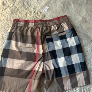 Rutig Burberry badshorts med snörning - Snygga badshorts kommer med påse från Burberry med klassiskt rutigt mönster i beige, svart, vitt och rött. Shortsen har elastisk midja med svart snörning och praktiska fickor. Perfekta för sommaren och strandhäng, med exklusiv känsla och ikonisk design.