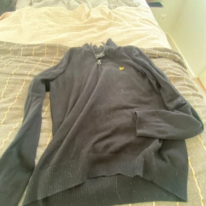 Svart half zip tröja från Lyle & Scott - Svart långärmad tröja från Lyle & Scott med ribbad krage och half zip-dragkedja. Tröjan har det klassiska gula logobroderiet på bröstet och är tillverkad i mjuk ullblandning. Perfekt för dig som gillar stilren och enkel design.