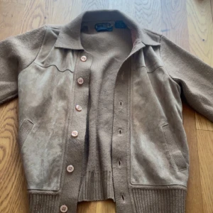 Beige cardigan jacka från Facade Club - Säljer en beige cardigan jacka från Facade Club i storlek M. Jackan har stickade partier och detaljer i äkta skinn framtill, med knappar och två fickor. Perfekt för dig som gillar vintagevibbar och snygga materialmixar.