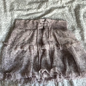 Blommig volangkjol från STR - Supersöt kort kjol från STR i ljuslila med små blommor. Kjolen har volanger, smock i midjan och knytband framtill. Perfekt för vår och sommar.