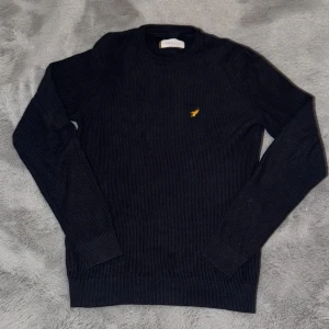 Mörkblå stickad tröja från Pier One - Snygg mörkblå stickad tröja från Pier One med rund halsringning och ett gult broderat fågelmärke på bröstet. Tröjan har långärmad passform och är perfekt för lager-på-lager-stil under kyliga dagar.