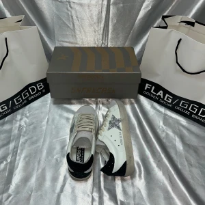 Golden Goose sneakers med glitterstjärna - Snygga vita sneakers från Golden Goose med silverglittrig stjärna på sidan och svart glitterdetalj på hälen. Skorna har beige mockadetaljer vid tån, klassisk snörning och platt sula. Perfekta för dig som vill sticka ut med en cool och trendig look.