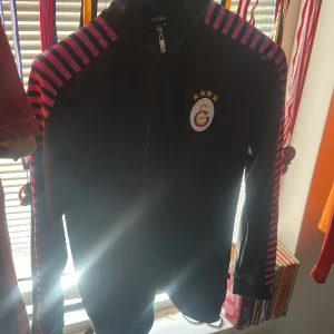 Svart randig jacka från GS Store - Svart jacka från GS Store med Galatasaray-logga på bröstet. Snygga rosa och röda ränder längs axlar och ärmar. Jackan har dragkedja framtill och är långärmad. Perfekt för dig som vill sticka ut med sportig stil.