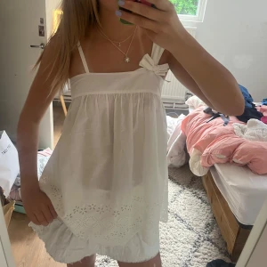 Vit blus med rosett och broderi - Supersöt vit blus med smala axelband och en stor rosett på ena axeln. Toppen är luftig och har broderade blommor längs nederkanten. Perfekt för varma dagar och ger en romantisk vibe. Tillverkad i bomull för en skön känsla.