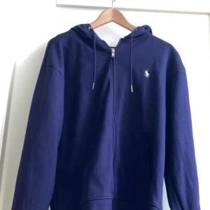 Marinblå hoodie från Polo Ralph Lauren - Snygg marinblå hoodie från Polo Ralph Lauren med dragkedja och huva. Klassisk logga broderad på bröstet och praktiska fickor framtill. Perfekt för en chill och stilren look. Tillverkad i mjuk bomull för extra komfort.