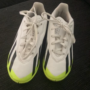 Adidas vita och neongula fotbollsskor - Snygga inneskor från Adidas i vitt med svarta ränder och neongula detaljer. Skorna har klassisk snörning och är tillverkade i syntetmaterial. Perfekta för dig som vill sticka ut på planen med stilrena och sportiga skor. 