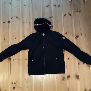 Svart Moncler Windbreaker  - Säljer en svart vindjacka från Moncler med huva och dragkedja framtill. Storler 1 i moncler vilket motsvarar S. Jackan har Moncler-logga på ärmen och snygga ränder på huvan. Perfekt för dig som vill ha en stilren och sportig look. Långärmad och med praktiska fickor. Denna har en liten repa på insidan (SYNS EJ FRÅN UTSIDAN) men därav priset ändå. Se sista bilden. Kontakta gärna vi minsta fundering.