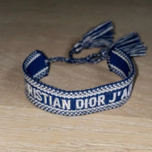 Blått Christian Dior J'Adior armband - Snyggt vävt armband från Christian Dior i blått och vitt med texten 'CHRISTIAN DIOR J'ADIOR'. Armbandet har justerbar knytning med tofsar i ändarna. Perfekt accessoar för att lyfta din outfit med en lyxig touch.