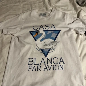 Casablanca T-shirt med svanmotiv - Kvitto finns vit  t-shirt från Casablanca med tryck av en svan och blå triangel på bröstet. Stora bokstäver 'CASA BLANCA PAR AVION' framtill. Klassisk rund hals och korta ärmar. Perfekt för dig som gillar stilrena statement-plagg.