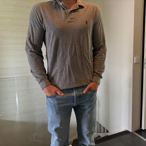 Grå långärmad pikétröja Polo Ralph Lauren - Snygg grå långärmad pikétröja från Polo Ralph Lauren med klassisk krage och knäppning framtill. Tröjan har en broderad logga på bröstet och är gjord i mjuk bomull. Perfekt för en clean och stilren look.