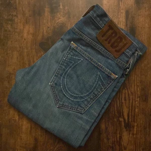 True Religion Jeans - Snygga jeans med slitningar och avklippta ändar för en rå look. Storlek 29. Skönt tvättade med en perfekt vintagekänsla. Passar till allt – sneakers, boots eller casual jacka. I fint skick trots slitage som hör till stilen.