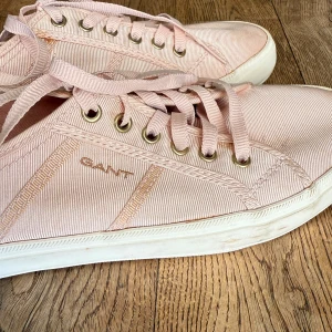 Ljusrosa sneakers från Gant - Snygga ljusrosa sneakers från Gant med klassisk design. Skorna har snörning, vita sulor och är tillverkade i bomullstyg med diskreta ränder. Perfekta för dig som gillar en stilren och fräsch look med en touch av pastell.