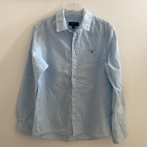 Ljusblå skjorta från GANT - Klassisk ljusblå skjorta från GANT med broderad logga på bröstet. Skjortan har lång ärm, knappar framtill och krage. Tillverkad i mjuk bomull som känns skön mot huden. Perfekt för en clean och stilren look.