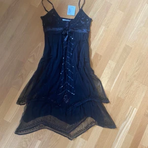 Svart glittrig klänning från POPIN - Supersnygg svart långklänning från POPIN med tunna axelband, v-ringning och glittriga paljettdetaljer. Klänningen har ett lager av transparent tyg, broderier, rosett framtill och asymmetrisk volangkant. Perfekt för dig som vill sticka ut med en elegant look. Storlek M men skulle snarare säga s
