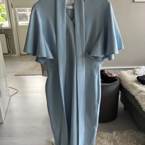 Ljusblå omlottklänning - Supersnygg ljusblå klänning med omlott och v-ringning. Klänningen har volangärmar, löstagbart knytband i midjan och en smickrande, figurnära passform som slutar vid knäna. Perfekt för dig som vill ha en stilren och feminin look. Använd fåtal gånger så i mycket fint skick!