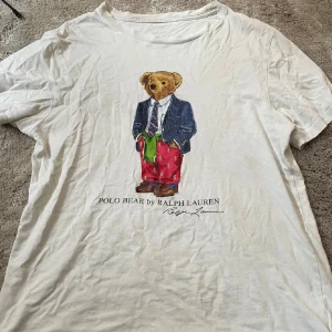 Vit Polo Bear t-shirt från Ralph Lauren - Vit t-shirt från Ralph Lauren med ikoniskt Polo Bear-tryck framtill. Klassisk rund hals och korta ärmar. T-shirten är i bomull och har en avslappnad passform, perfekt för en chill och stilren look.