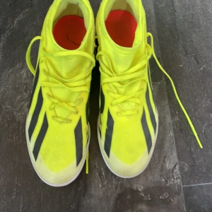 Neongula Adidas fotbollsskor - Snygga neongula fotbollsskor från Adidas med svarta ränder och vita sulor. Skorna har snörning och en strumpliknande passform för extra komfort. Perfekta för dig som vill sticka ut på planen och ha grym kontroll på bollen.