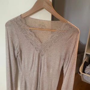 Beige långärmad topp med spets från intimissimi - Superfin beige långärmad topp från intimissimi med v-ringning och bred spetskant runt halsen. Mjuk och stretchig bomullsblandning som sitter snyggt på kroppen. Perfekt att styla med jeans eller kjol för en chill look.