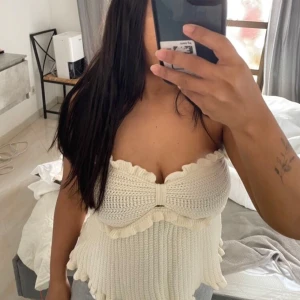 Stickad bandeau tubtopp i vitt - Supersöt stickad bandeau tubtopp i vit färg med volangdetaljer upptill och nedtill. Toppen är ärmlös och har en ribbad struktur som ger en snygg och trendig look. Perfekt för varma dagar när du vill ha något luftigt och stilrent.