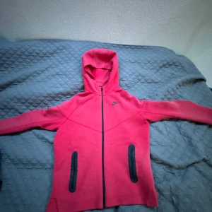 Rosa hoodiejacka från Nike XL - Nike hoodiejacka i stark rosa med svarta detaljer och dragkedja framtill. Jackan har huva, två fickor med svarta dragkedjor och är långärmad. Perfekt för dig som gillar sportig och chill stil. Äkta!!!
